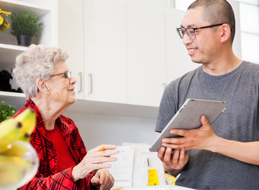 An intervenor holds up a tablet with large print notes for an older client to read. They are in a kitchen together.