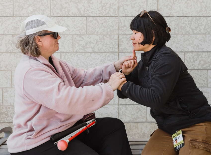 An intervenor uses tactile ASL to communicate with a person who is Deafblind. They are sitting together on a bench.